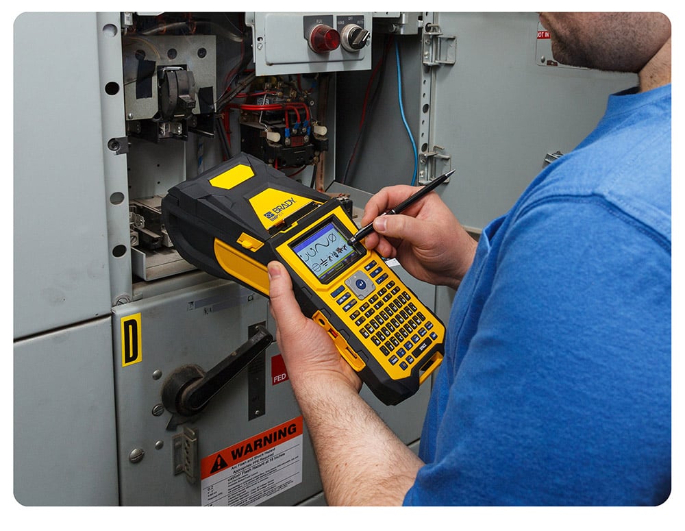 Maintenance tech holding a Brady BMP61 stands next to equipment.