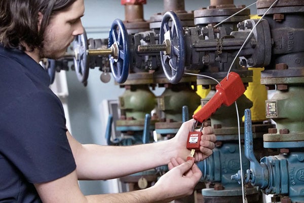 Man securing a red cable lockout device onto industrial pipes and valves.