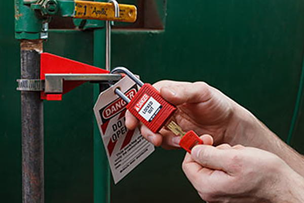 Hands attaching a lockout padlock and tag to a valve lockout device on industrial piping.