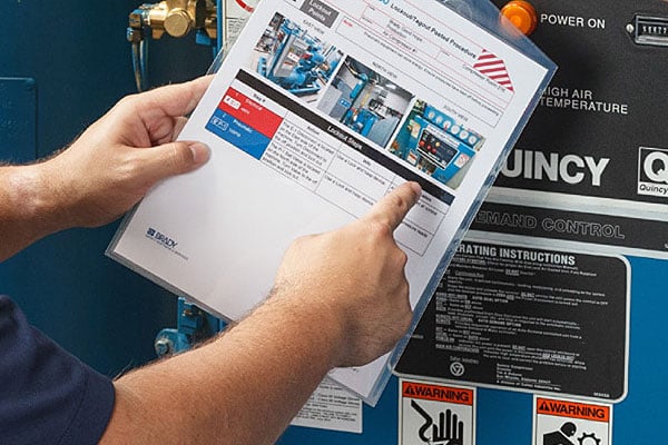 Hands point to the "Lockout Steps" on a plastic-sleeved Lockout/Tagout (LOTO) procedure document, posted on blue industrial equipment.