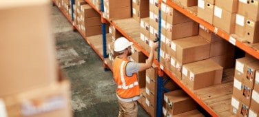 Worker in hardhat scans shelved packages in warehouse setting. 