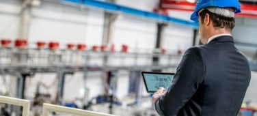 Man in suit and hardhat uses tablet in a factory.