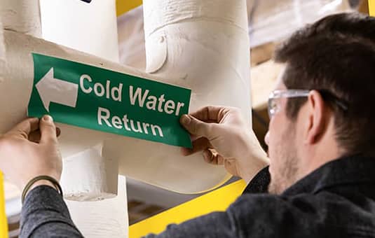 Employee applying a cold water return label to an overhead pipe.