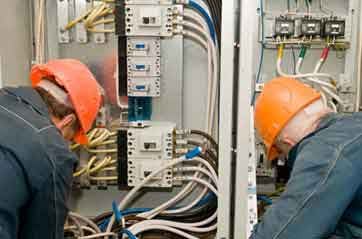 Two men in hard hats address a panel of wires.
