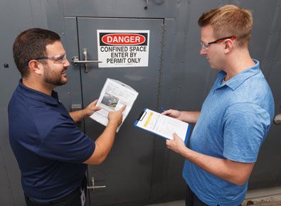 Two men reviewing paperwork in front of a confined space entry door.