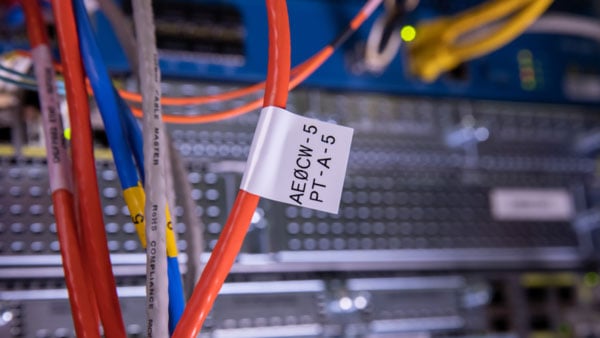 A close-up shot shows a white Brady QuickFlag label to a bundle of network cables in a data center. The label is designed to wrap around the cables and has a printed identifier.