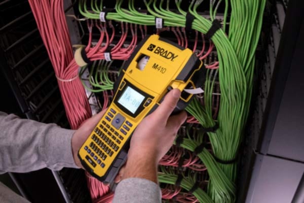 A person's hand holds a black and yellow portable Brady M410 label printer in a data center environment. The printer has a label partially dispensed, showing printed text intended for cable identification.