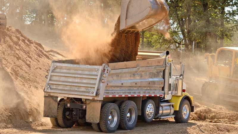 A dump truck being filled with dirt by an excavator.