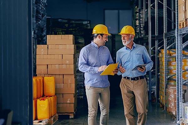 Two men in hardhats in a warehouse have a conversation.