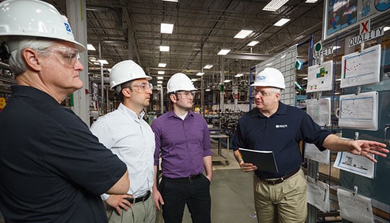 Four men in hardhats in an industrial setting discuss topics.