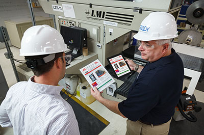 Two men in hardhats discuss procedures, holding printed process sheet.