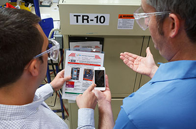 Two men in safety goggles discuss LOTO procedures, referencing a sheet.