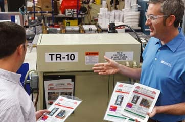 Two men discuss industrial equipment while holding brochures.