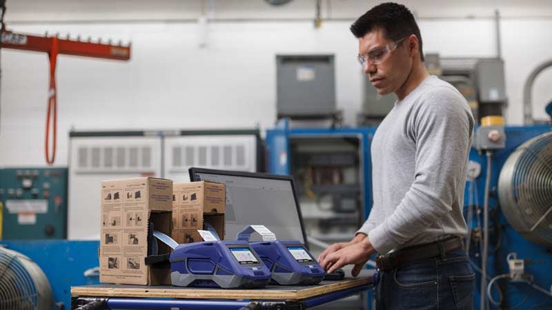 A worker stands next to two blue label printers.
