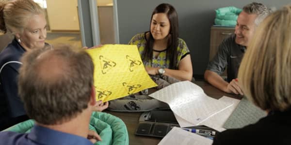 Workers sit at a table, inspecting a yellow warning label.