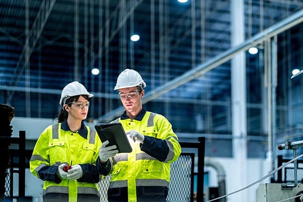 Two workers in safety gear review processes on a tablet.
