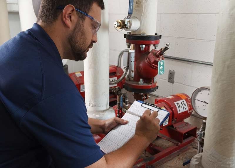 Worker inspects pumps and valves, writes on a clipboard.