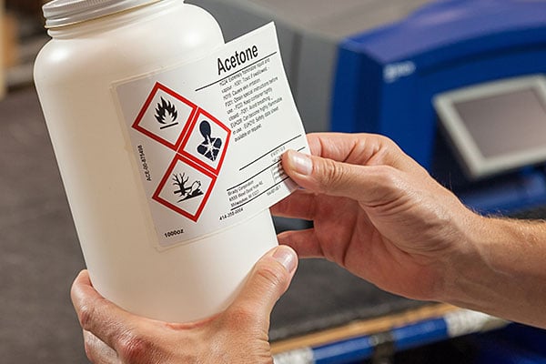 Hands label a white bottle with a GHS warning.