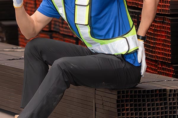 Worker in safety vest sits with hand on lower back.