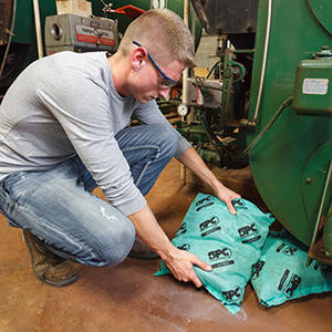 Man kneels to place absorbent pillows under industrial machinery.