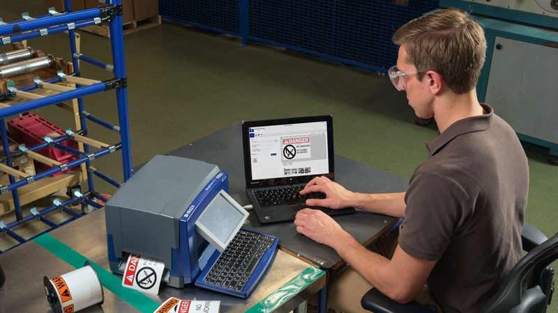 A man works on a laptop, with a label printer nearby.
