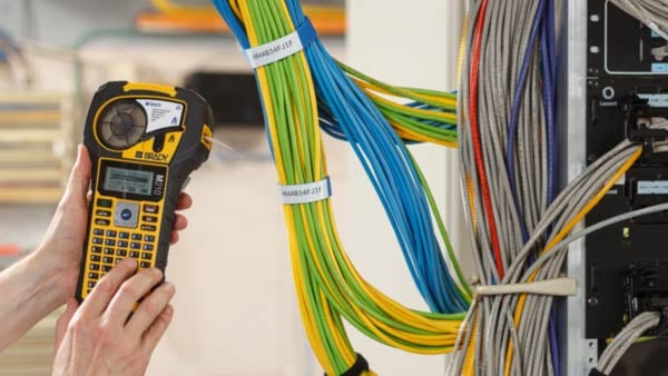 Datacom worker holding a Brady M210 printer in front of a server rack with data cables of varying colors.