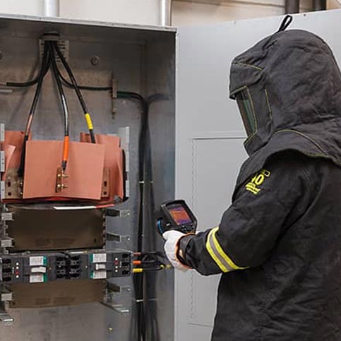 Worker in heavy PPE in front of electrical panel.