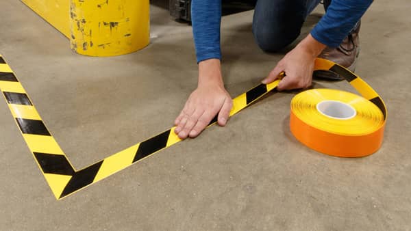 Close-up of a person&#x27;s hands applying yellow and black striped hazard floor marking tape in on a concrete floor, with the roll of tape visible to the right.