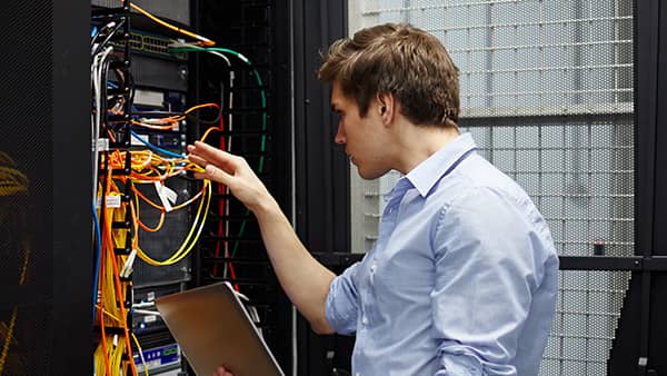 A man  working in an open server rack in a data center, configuring network cables with one hand while holding a silver laptop in the other.