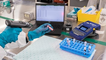 Lab technician scanning vial with laptop and printer in background. 